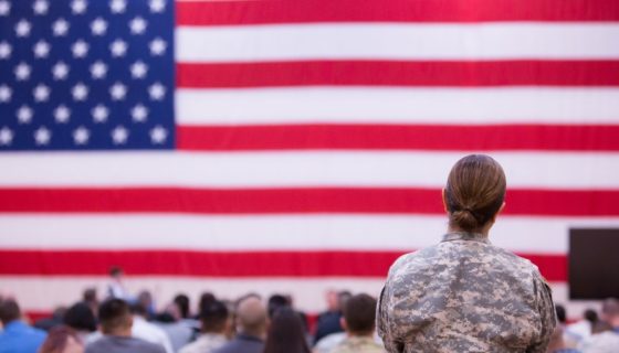 Service member in an audience looking in the direction of a large US flag.