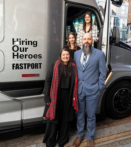 2023 Transition Trucking contest winner poses for a photo with his wife and two daughters in front of the Kenworth truck featuring the Hiring Our Heroes and Fastport logos.