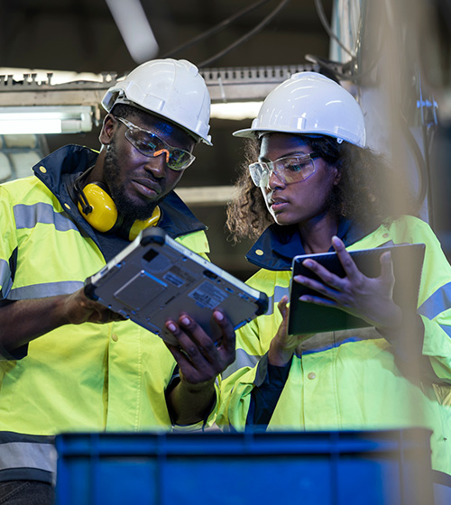 male and female workers wearing yellow reflective jackets and hard hats and holding tablets review information from one tablet.