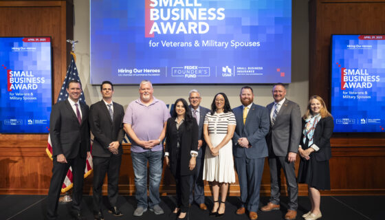 Hiring Our Heroes Small Business Grant Program Awards Celebration. A group of nine people standing in front of a large screen displaying "Small Business Award for Veterans & Military Spouses." The screen also features logos for Hiring Our Heroes, FedEx Founder's Fund, USAA, and Small Business Insurance. Two smaller screens on either side display the same award title and date, April 29, 2025. An American flag is visible to the left of the group.