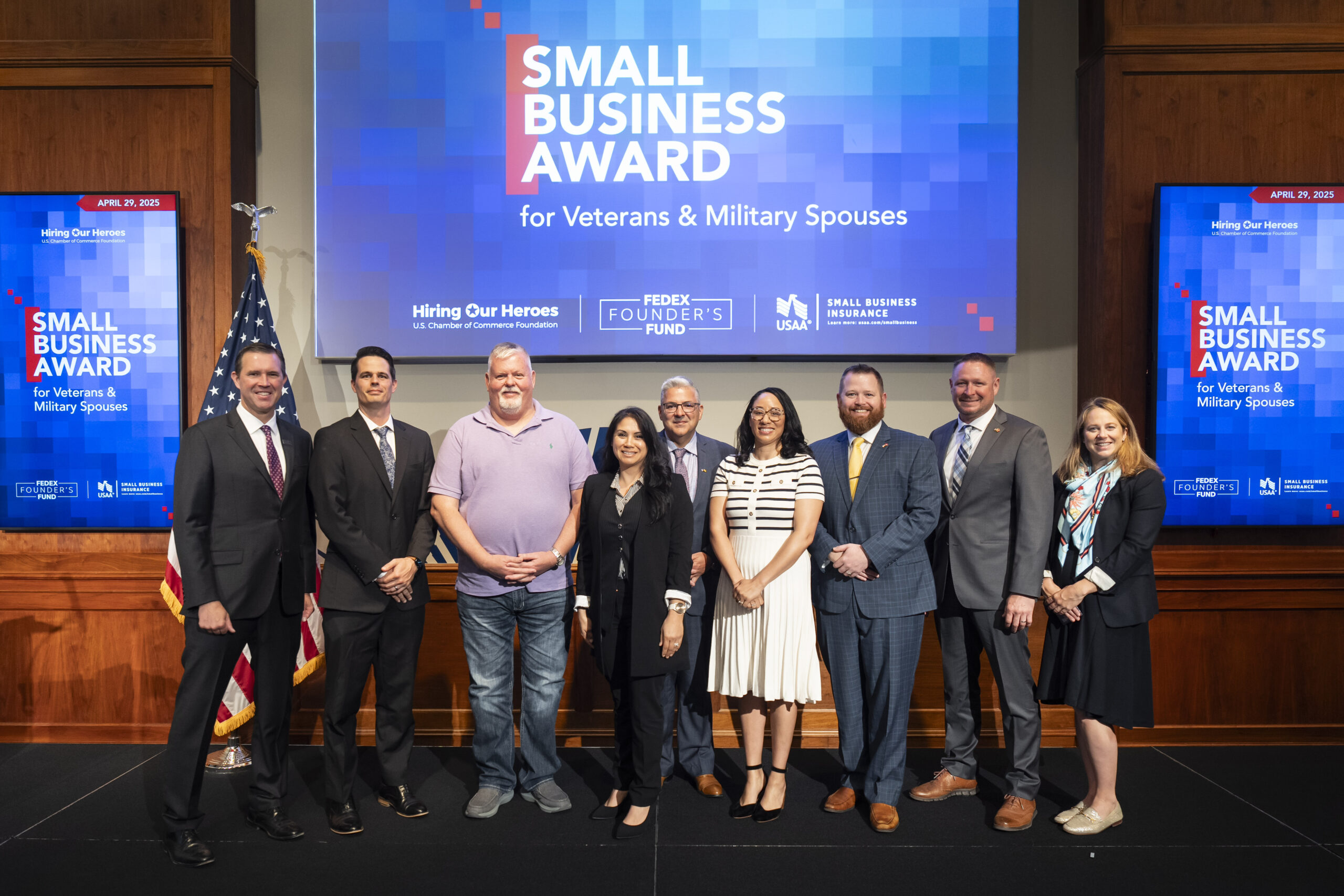 Hiring Our Heroes Small Business Grant Program Awards Celebration. A group of nine people standing in front of a large screen displaying "Small Business Award for Veterans & Military Spouses." The screen also features logos for Hiring Our Heroes, FedEx Founder's Fund, USAA, and Small Business Insurance. Two smaller screens on either side display the same award title and date, April 29, 2025. An American flag is visible to the left of the group.