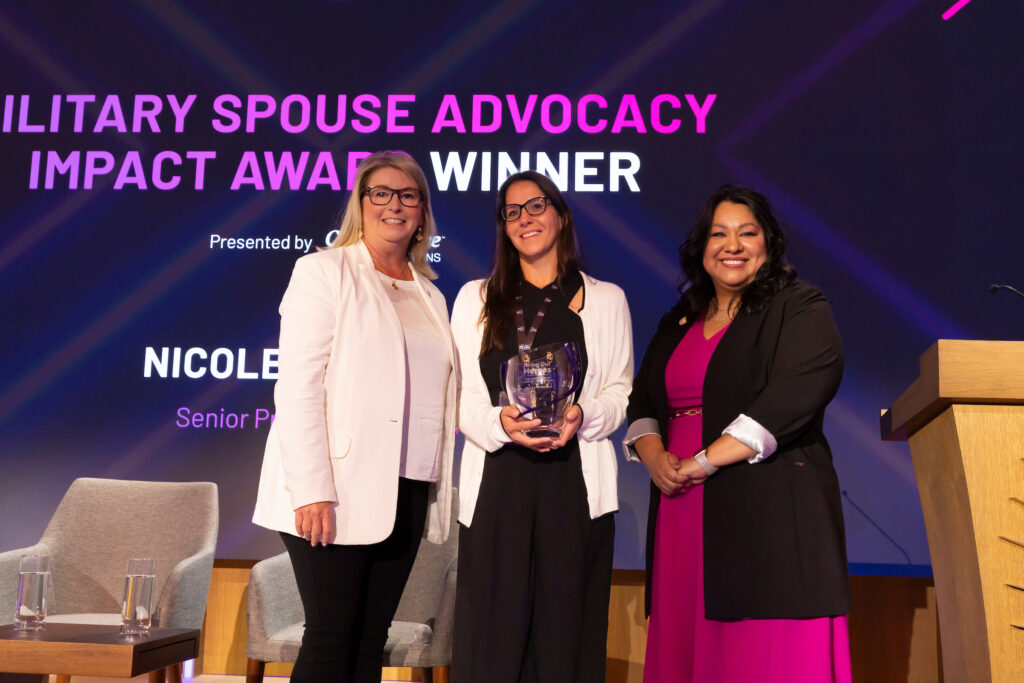 Three individuals are standing on a stage, with the middle person holding an award. The background displays text that reads 'MILITARY SPOUSE ADVOCACY IMPACT AWARD WINNER'