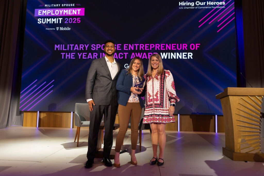 Three individuals stand on a stage in front of a large screen displaying the text 'Military Spouse Employment Summit 2025' and 'Military Spouse Entrepreneur of the Year Impact Award Winner.' The event is sponsored by Hiring Our Heroes and T-Mobile. The person in the center holds an award, while the other two stand on either side. The background features purple diagonal lines and logos for Hiring Our Heroes and U.S. Chamber of Commerce Foundation.
