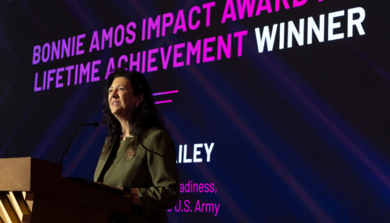A person stands at a podium on stage, with a large screen behind displaying the text 'BONNIE AMOS IMPACT AWARD FOR LIFETIME ACHIEVEMENT WINNER' in bold white and pink letters. The event is titled 'EMPOWERING WOMEN SUMMIT 2023' and is sponsored by T-Mobile. The name 'RILEY' and the title 'Director of Readiness, U.S. Army' are also visible on the screen.