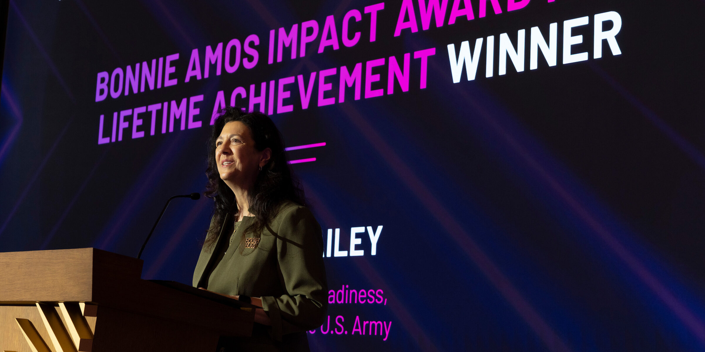 A person stands at a podium on stage, with a large screen behind displaying the text 'BONNIE AMOS IMPACT AWARD FOR LIFETIME ACHIEVEMENT WINNER' in bold white and pink letters. The event is titled 'EMPOWERING WOMEN SUMMIT 2023' and is sponsored by T-Mobile. The name 'RILEY' and the title 'Director of Readiness, U.S. Army' are also visible on the screen.