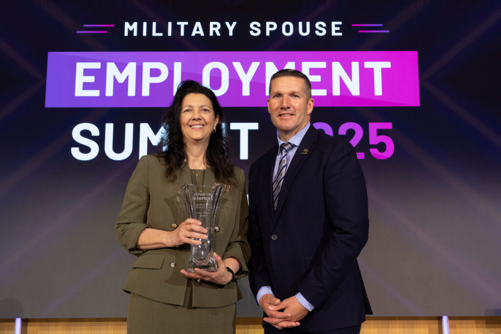 Two individuals are standing side by side in front of a backdrop that reads 'MILITARY SPOUSE EMPLOYMENT SUMMIT 2025.' The individual on the left is holding a glass award.