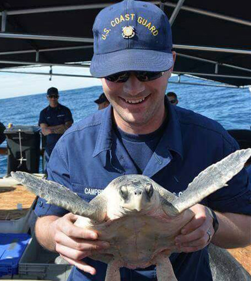 Rhys Campbell, a transitioning member of the U.S. Coast Guard, smiles while holding a sea turtle who is less than impressed with having his photo taken.
