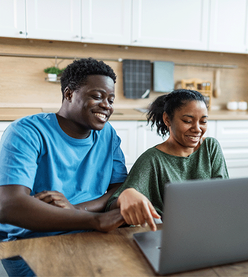 Photo of a couple smiling while working on a laptop at a kitchen table.