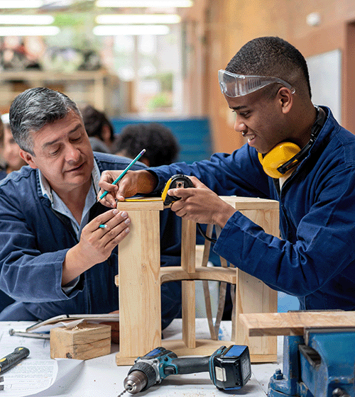 A shop teacher helps a student work on a wood project using a tape measure, hammer and a drill.