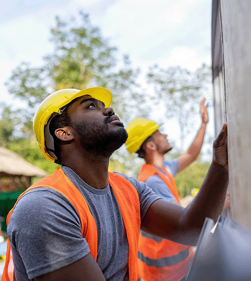 Two construction workers wearing yellow hard hats and orange vests work on putting up a wall.