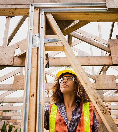 A female construction worker wearing a yellow hard hat and orange safety vest works on a wooden doorway.