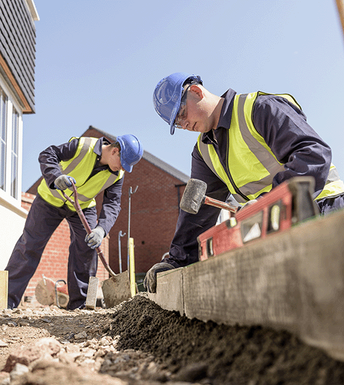 Two construction workers wearing blue hard hats and yellow safety gear work on a masonry project using a shovel and a mallet.