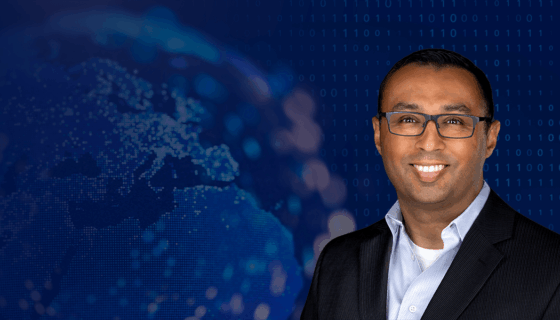 A man wearing glasses and a dark suit jacket over a light blue shirt is smiling against a dark blue background. The background features a blurred digital globe with illuminated dots and binary code patterns, symbolizing technology and connectivity.
