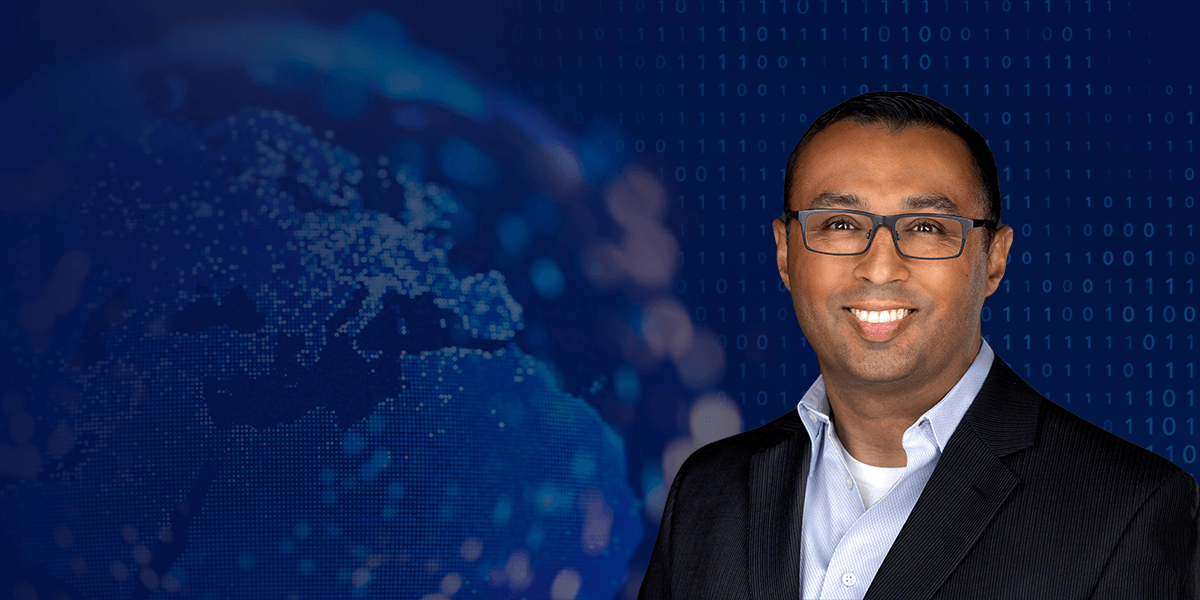 A man wearing glasses and a dark suit jacket over a light blue shirt is smiling against a dark blue background. The background features a blurred digital globe with illuminated dots and binary code patterns, symbolizing technology and connectivity.