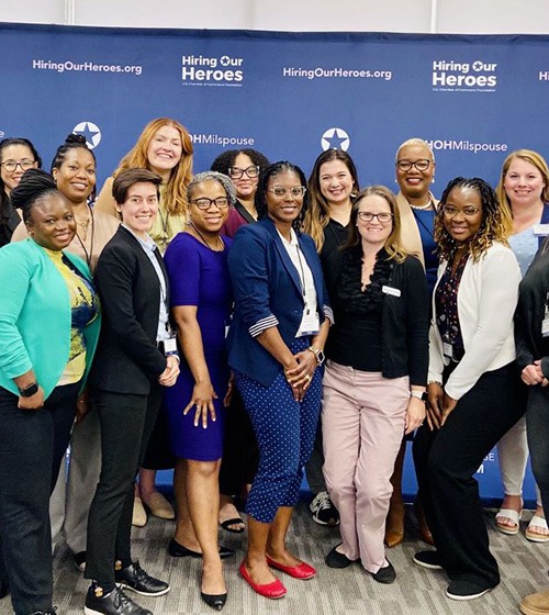 Women posing and smiling in front of a Hiring Our Heroes backdrop at an Amplify event.