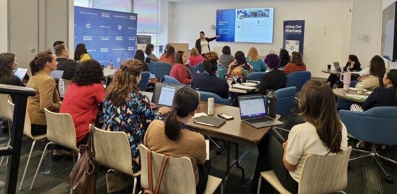 Attendees sitting at tables with laptops hear from presenter during an in-person Amplify