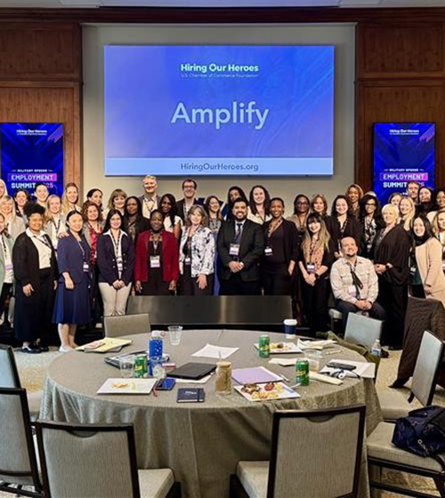 Amplify event attendees pose for a photo in front of a screen displaying a title slide for Hiring Our Heroes Amplify.