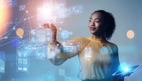 A woman in a yellow blouse stands against a blue background, holding a tablet in one hand while interacting with floating digital data interfaces and graphs, representing data visualization.