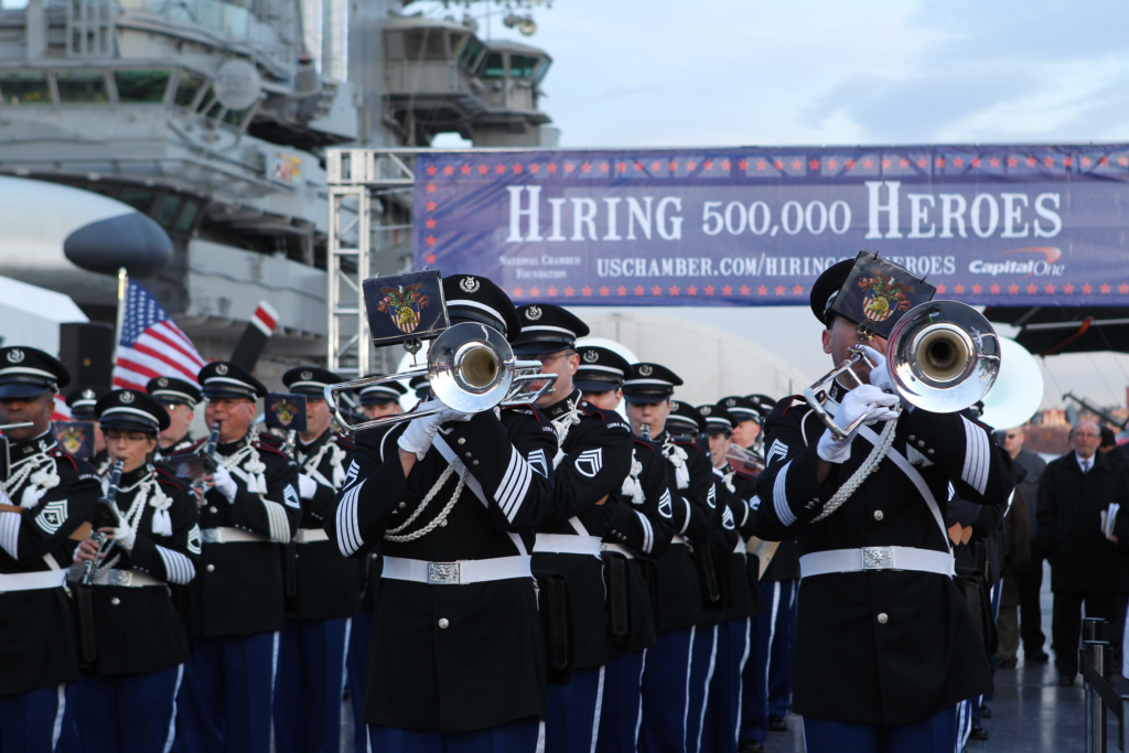 A marching band plays in front of a Hiring 500,000 Heroes banner.