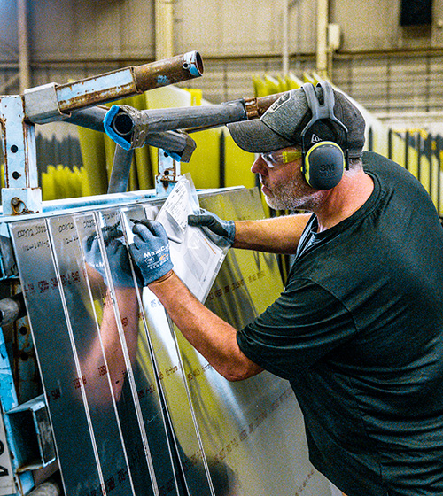 Man wearing a hat, ear and eye protection marking sheet metal in a manufacturing facility