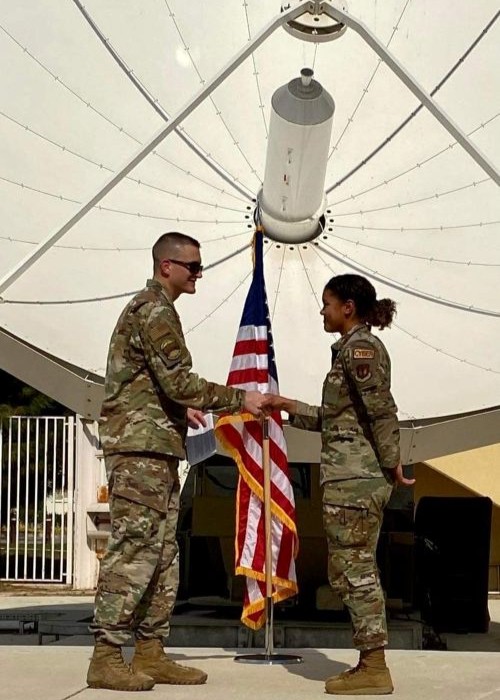Lyric Green in uniform shaking hands with another person in uniform standing in front of a flag and satellite dish.