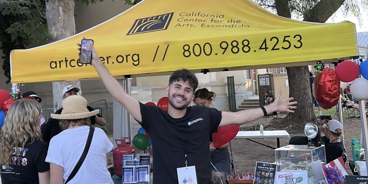 Brandon Hayes with his arms out wide in front of a yellow CCAE pop-up cover at a street fair.