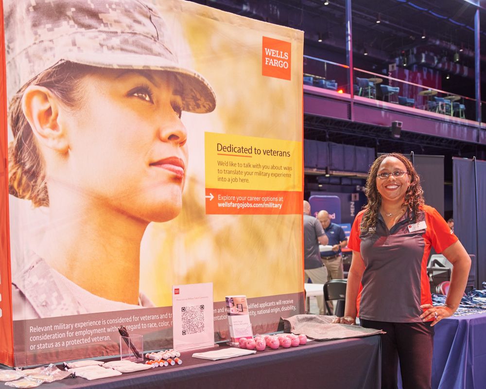 Hiring fair employer participant poses in front of her booth that features a large image of a female service member.