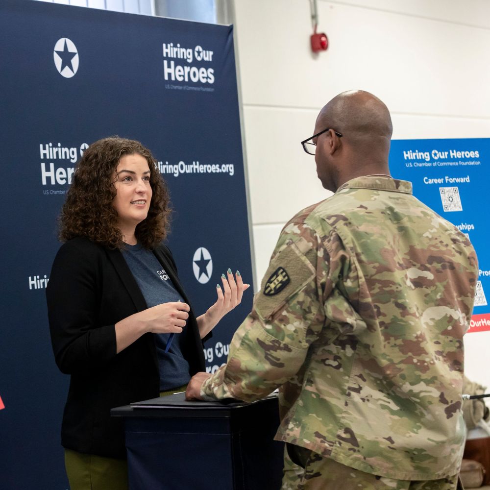 A woman talks to a man in uniform in front of a Hiring Our Heroes blue backdrop.