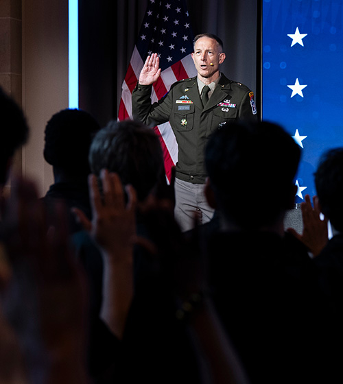 A military leader in uniform holds up his right hand in front of a crowd of people also holding up their right hands.