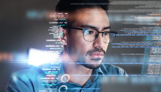 A serious man with glasses looks at a computer with data superimposed over the image.