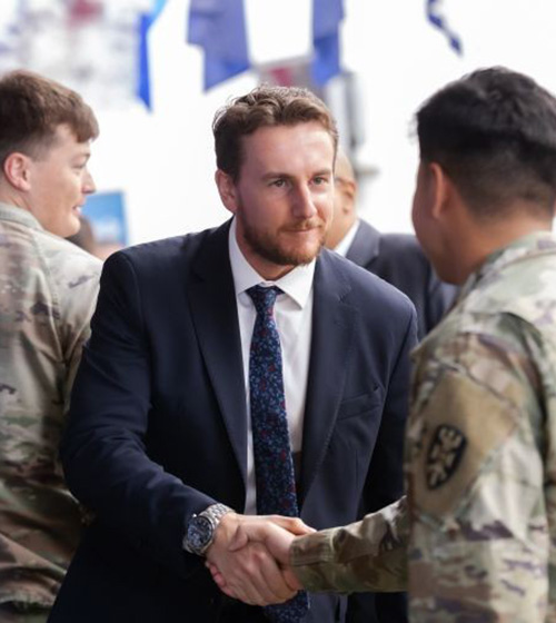 Two men, one in a suit, and the other in a military uniform, shake hands and make eye contact at a hiring event.