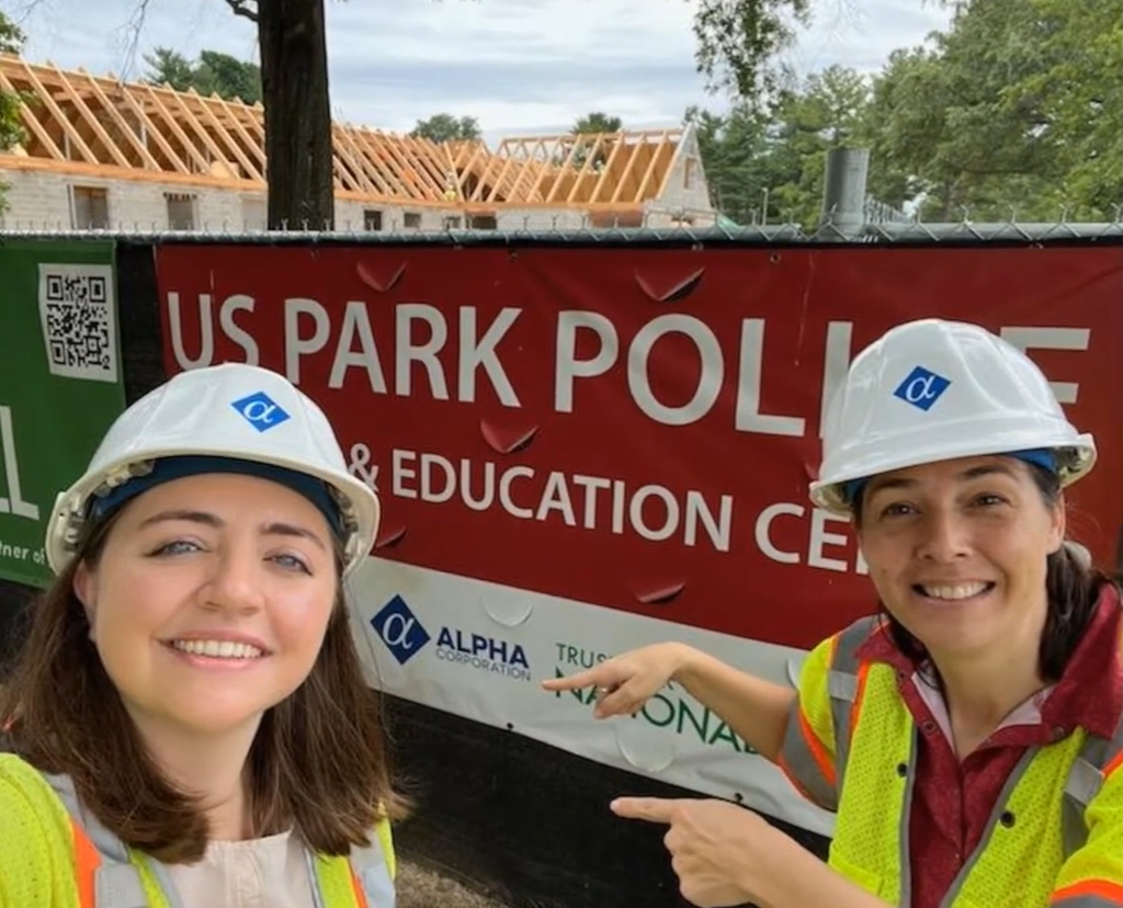 Two women fellows with hard hats stand in front of a sign with their host company's logo on it.