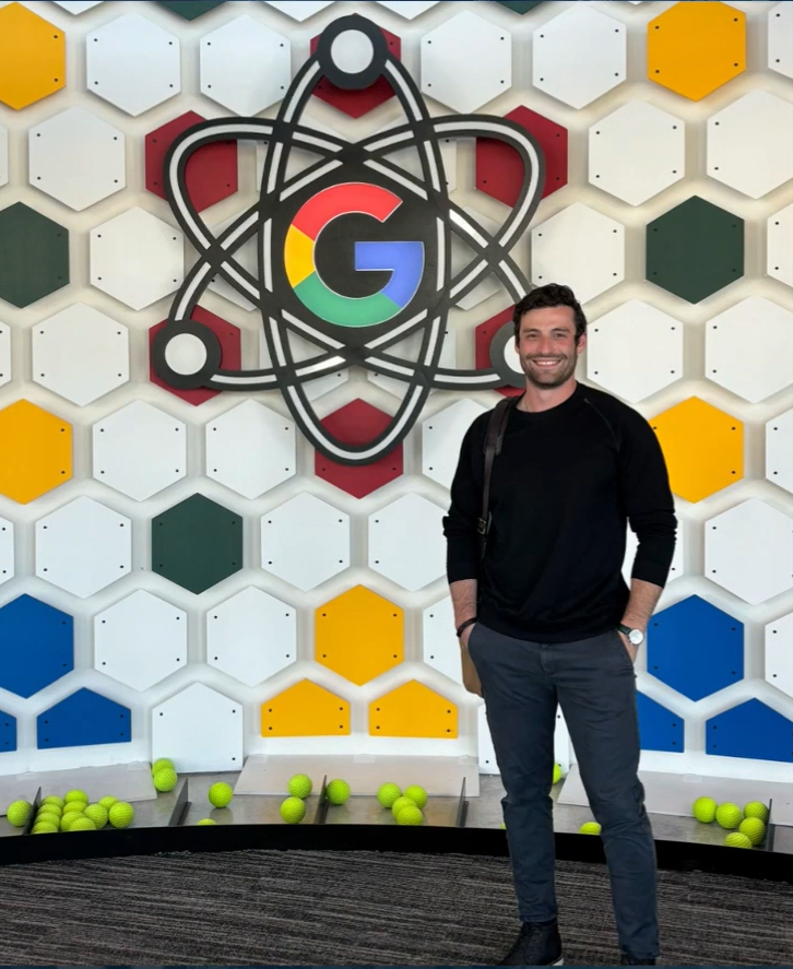 A fellow from 2024 poses in front of the Google 'G' logo inside of an atom shaped piece of art.