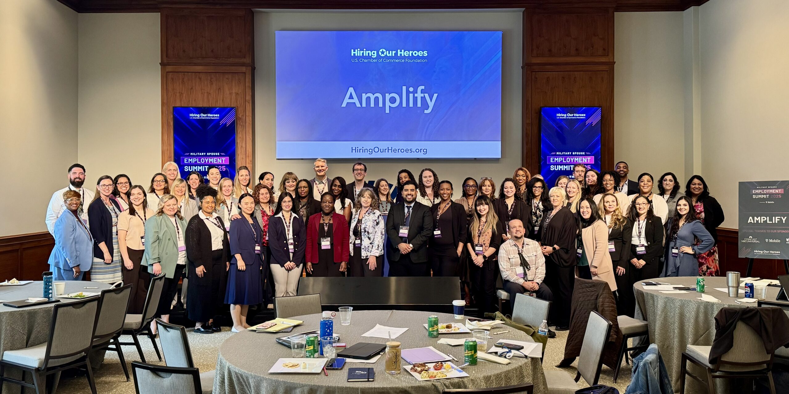 A group of military spouses posing under a monitor with the word Amplify on it.