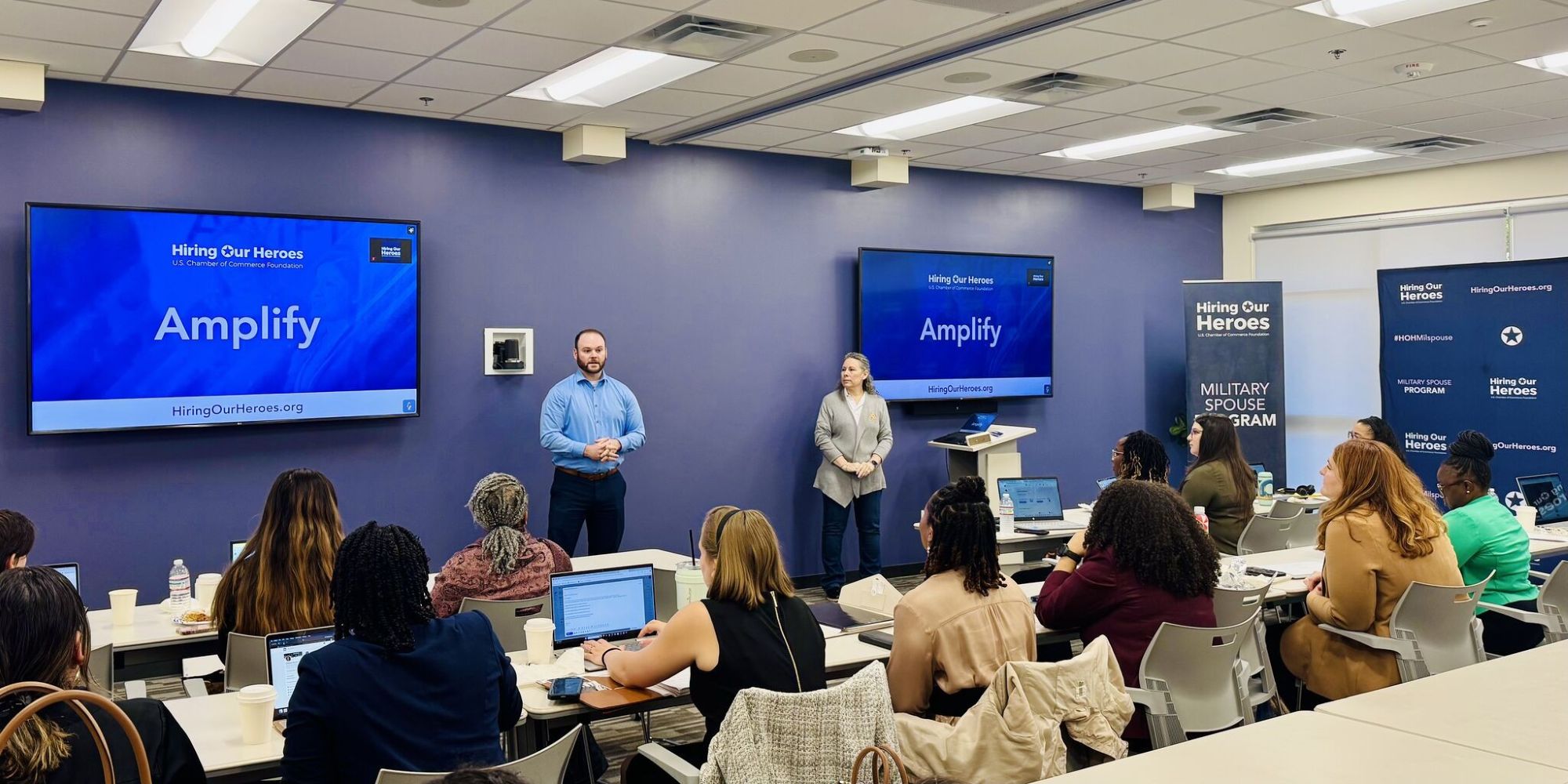 A group of individuals sit at tables with computers and laptops while listening to two presenters at the front of the room. Monitors display Amplify signage.